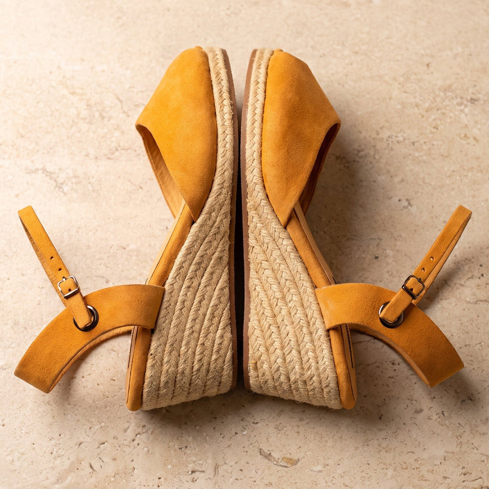 A pair of handcrafted leather ochre wedge espadrille sandals with jute sole by Miriam Bella with adjustable ankle straps and braided jute wedge heels, displayed on a textured beige stone surface.