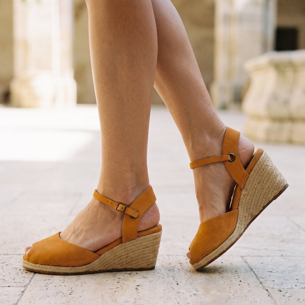 A person wearing tan leather wedge sandals with espadrille-style woven soles, shown from the knees down on a light stone walkway.