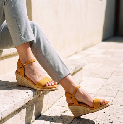 Woman wearing Gaia ochre leather open toe espadrille wedge sandals with grey pants standing on a stone terrace