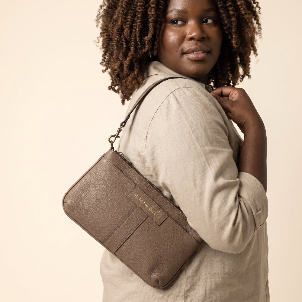 A woman wearing a light beige linen shirt carries a taupe brown pebbled leather mini purse with a gold-tone logo plate that reads 'Miriam Bella'. The photo is taken against a solid, warm beige background.