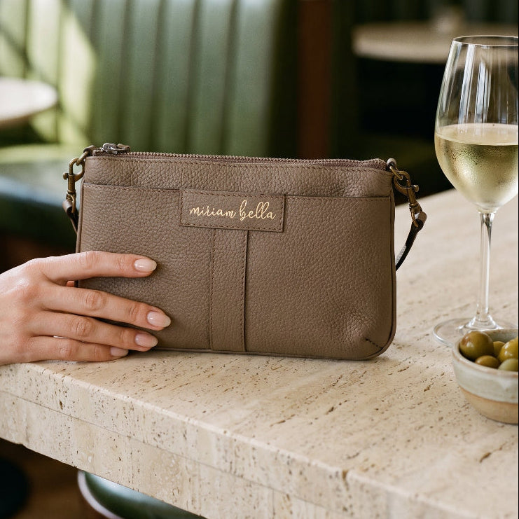 A hand holding the Mini Tote in Moss colour on table in a cocktail bar with a glass of white wine and olives on the table.