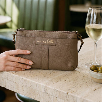 A hand holding the Mini Tote in Moss colour on table in a cocktail bar with a glass of white wine and olives on the table.