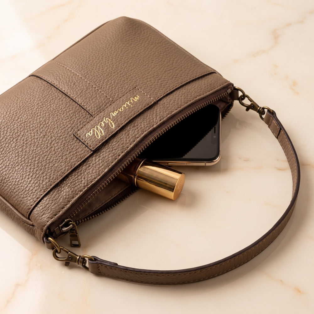 A brown pebbled leather mini purse with a gold script logo patch, shown open on a light marble surface with a smartphone and gold lipstick inside.