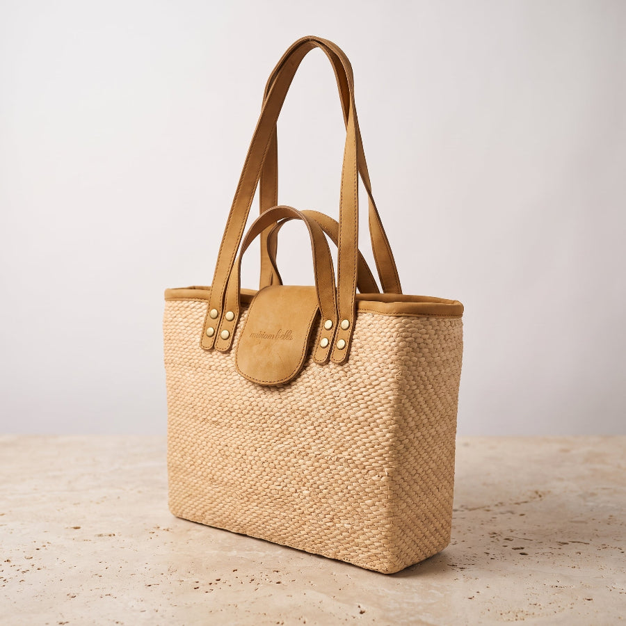 A woven straw tote bag with tan leather handles and a matching leather flap closure, standing on a light stone surface against a plain light gray background.