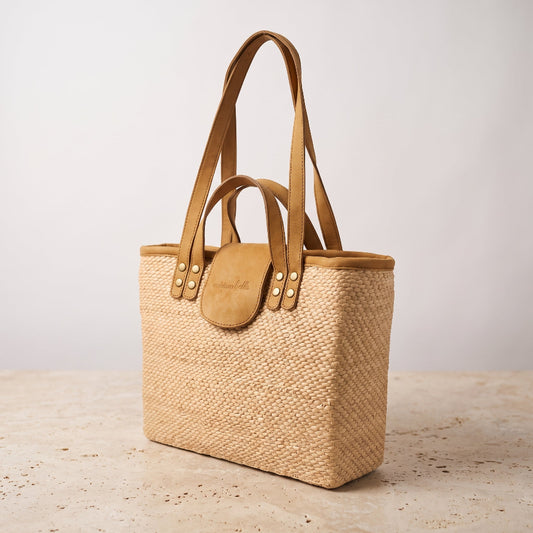 A woven straw tote bag with tan leather handles and a matching leather flap closure, standing on a light stone surface against a plain light gray background.