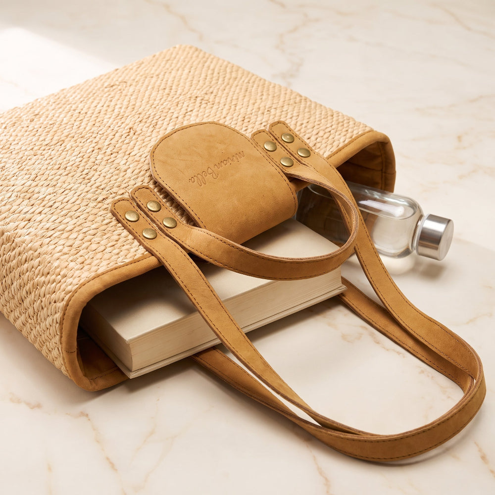 A woven straw tote bag with tan leather handles and trim, styled on a light marble surface with a book and a small glass bottle inside.