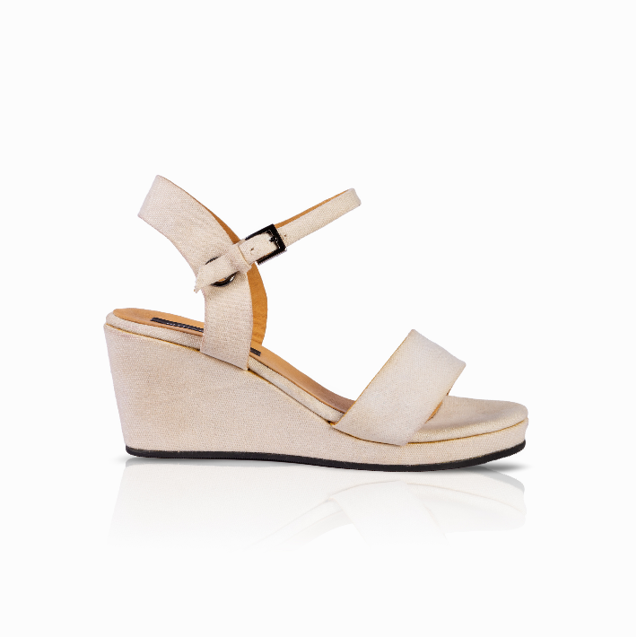 A single women's wedge sandal in a light beige or cream color, featuring a wide front strap, a slim ankle strap with a small black buckle, and a wedge heel, shown against a plain white background with a soft reflection.