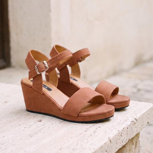 A pair of brown wedge sandals with ankle straps and silver buckles, resting on a light stone ledge against a blurred beige wall background.