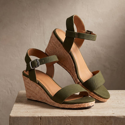A pair of olive green wedge sandals with wide straps, ankle buckles displayed on a textured stone pedestal against a neutral studio background.