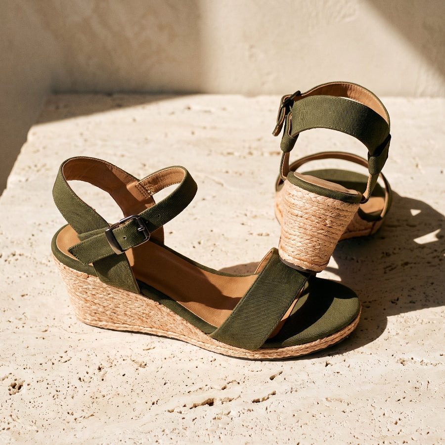 A pair of wedge sandals with olive green straps and a natural jute-wrapped heel, displayed on a textured beige stone surface in warm sunlight.