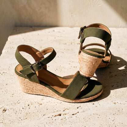 A pair of wedge sandals with olive green straps and a natural jute-wrapped heel, displayed on a textured beige stone surface in warm sunlight.