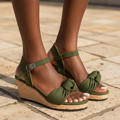 Olive green wedge sandals with ankle straps and knotted front details, shown on a model's feet against a light gray tiled floor.