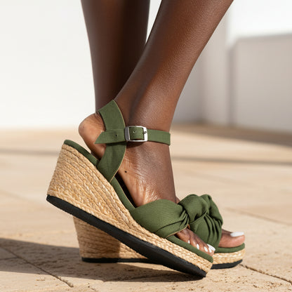 Olive green wedge sandals with a knotted bow detail on the front and an adjustable ankle strap, shown on a model's feet against a light gray tiled floor.