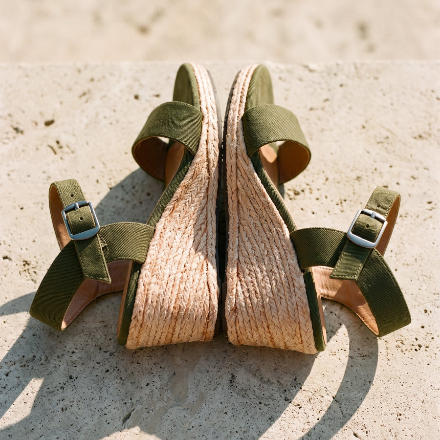 A pair of women's wedge sandals with olive green straps and woven jute wedge heels, photographed on a light stone surface outdoors.