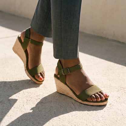 Woman in a gray trouders and olive classic wedge espadrille sandals walking on a stone terrace.