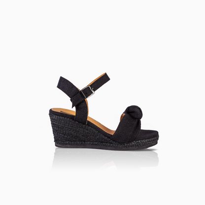 A single black wedge sandal with an ankle strap and a knotted detail on the toe, shown from a side profile against a white background with a soft reflection.