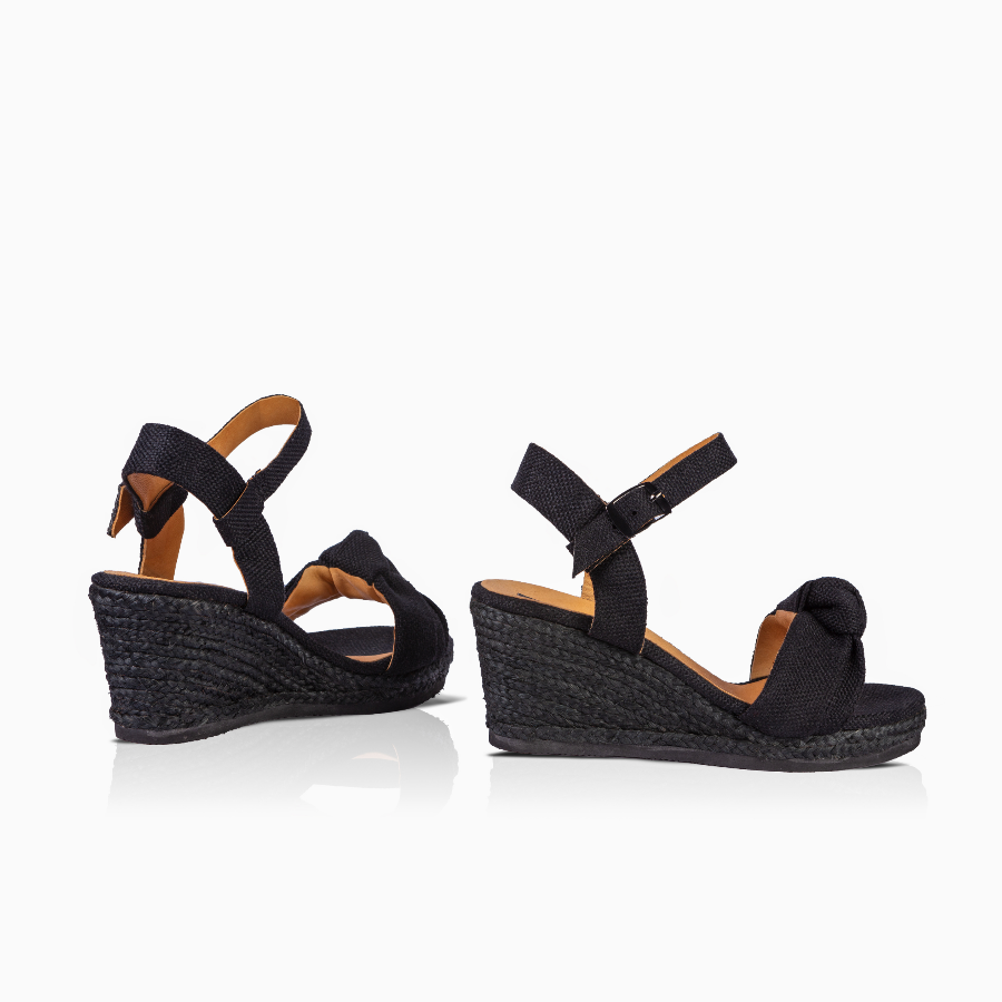 A pair of black wedge sandals with ankle straps and a knotted detail on the toe, shown from the side against a white background.