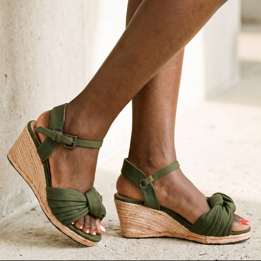 A person wearing olive green wedge sandals with ankle straps and braided toe detail, standing on a light stone floor next to a wall.