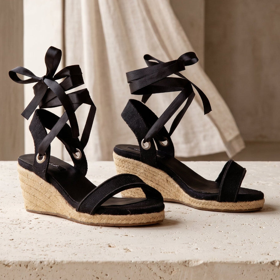 Close-up of Gaia black open toe canvas espadrille wedge heels with white ribbon ankle ties and jute wedge on a stone bench.