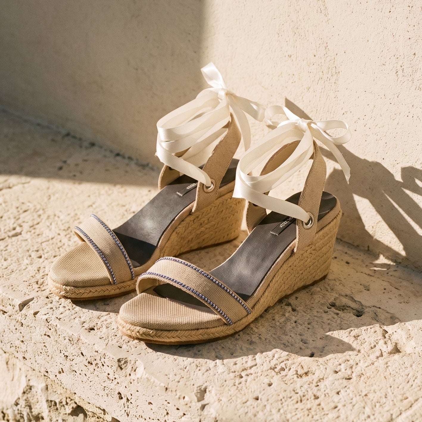 Close-up of Gaia cream open toe canvas espadrille wedge heels with cream ribbon ankle ties and jute wedge on a stone bench.