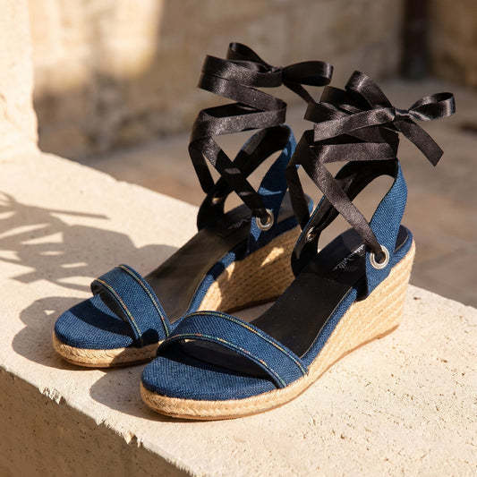 Close-up of Gaia indigo open toe canvas espadrille wedge heels with black ribbon ankle ties and jute wedge on a stone bench.