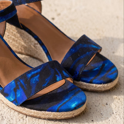 Close up of Blue and black patterned wedge sandals with espadrille-style jute-wrapped heels and ankle straps, photographed on a light stone surface.