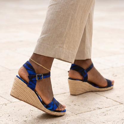 A close-up of a person wearing blue patterned wedge espadrille sandals with beige linen pants, walking on a light stone tiled pavement.