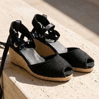 Close-up of Gaia black peep toe canvas espadrille wedge heels with white ribbon ankle ties and jute wedge on a stone bench.