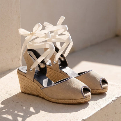 Close-up of Gaia peep toe cream canvas espadrille wedge heels with white ribbon ankle ties and jute wedge on a stone bench.