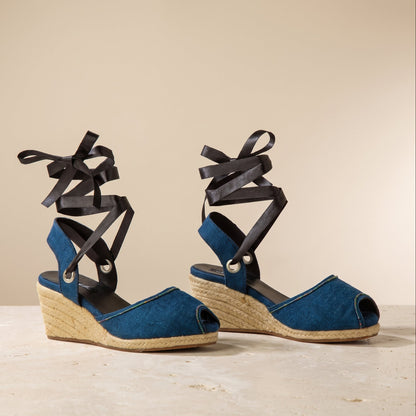 A pair of blue peep toes wedge sandals with black ribbon ankle straps, displayed on a light stone surface against a neutral beige background.