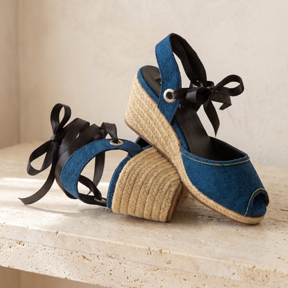 A pair of blue peep toes wedge sandals with black ribbon ankle straps, displayed on a light stone surface against a neutral beige background.