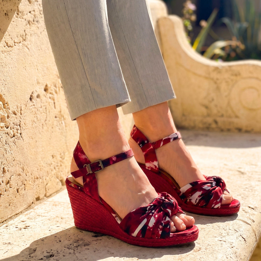 A person wearing red and burgundy floral wedge sandals with ankle straps and knotted front straps, seated on a stone bench outdoors.