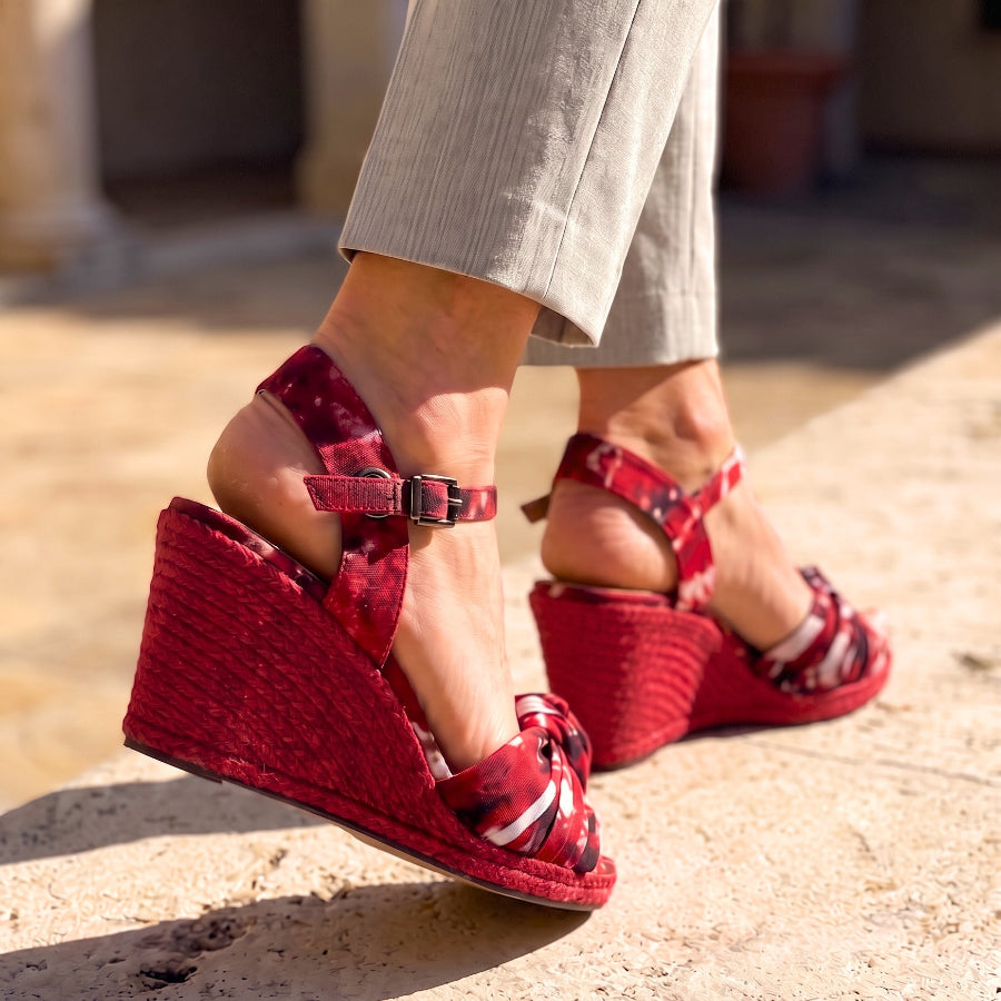 A close-up of a person wearing red patterned wedge sandals with ankle straps and beige trousers, standing on a sandy stone surface.