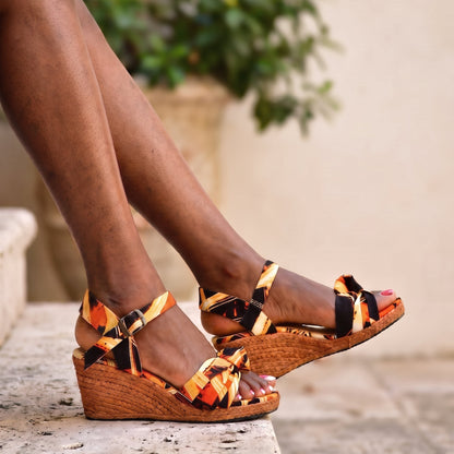 A person wearing wedge sandals with a colorful orange, black, and yellow patterned fabric upper and woven jute wedges, sitting on a stone ledge with a blurred background.