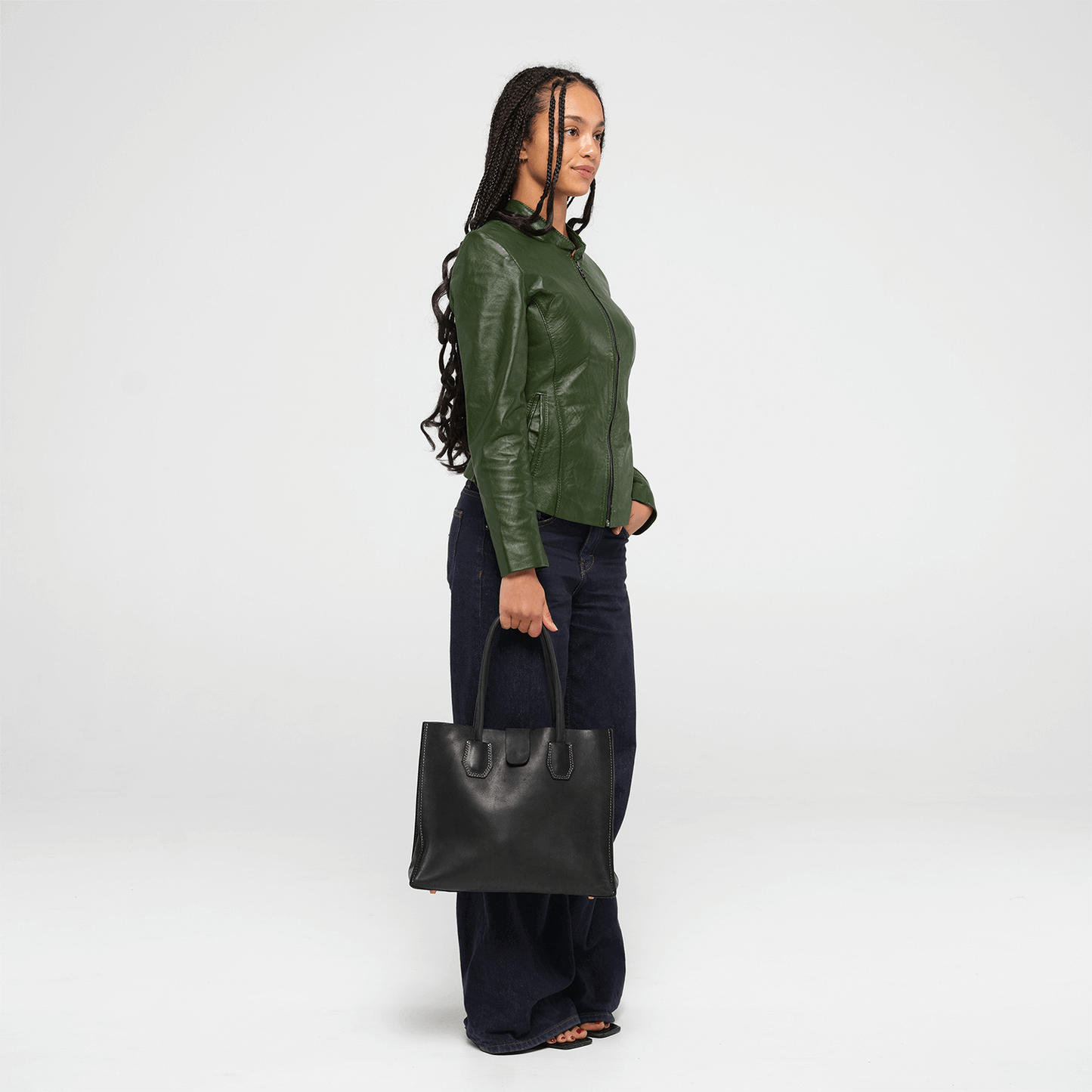 Person wearing a green leather jacket and holding a black leather tote bag on a white background