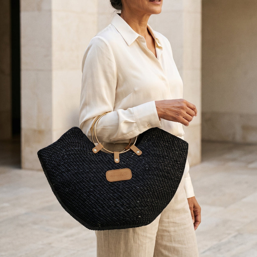 A woman in a cream outfit holding a black Jua woven straw beach bag in with brass ring handles.