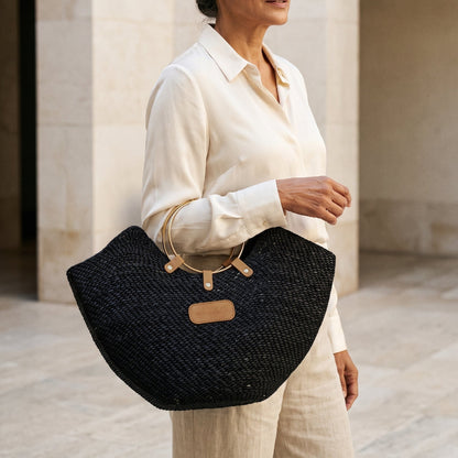 A woman in a cream outfit holding a black Jua woven straw beach bag in with brass ring handles.