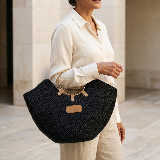 A woman in a cream outfit holding a black Jua woven straw beach bag in with brass ring handles.