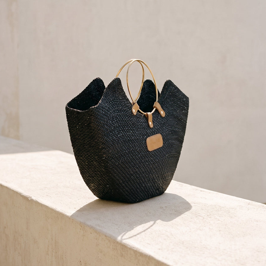 A black Jua woven straw beach bag with brass ring handles on a stone bench.