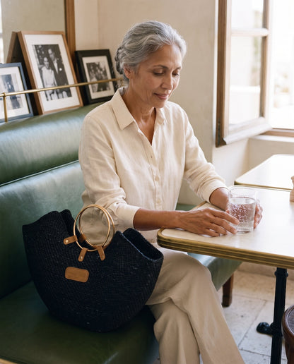 A woman in a cafe sitting next to Jua woven straw beach bag with brass ring handles in black.