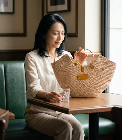 A woman in a cafe placing the Jua woven straw beach bag with brass ring handles in natural on a cafe table.