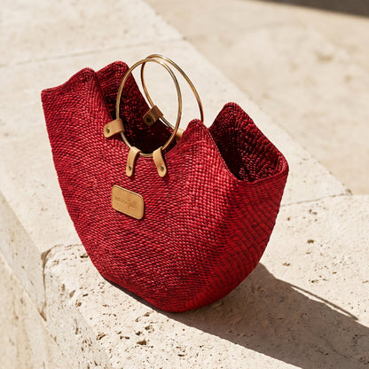 A red Jua woven straw beach bag with brass ring handles on a stone bench.