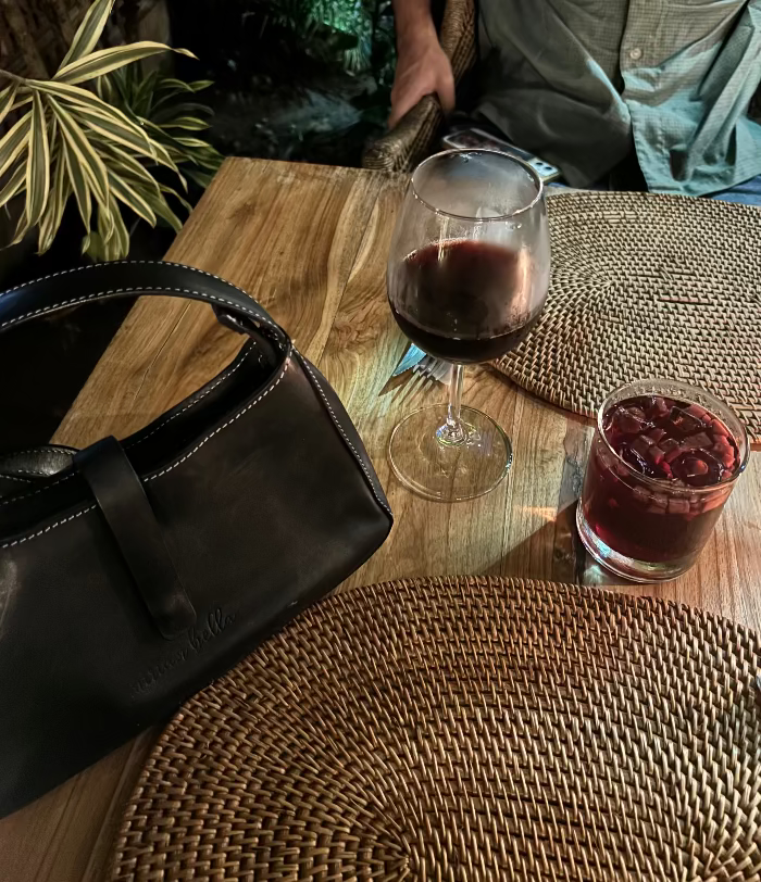 Two glasses of red wine on a wooden table with a black leather mini shoulder bag and a plant in the background.