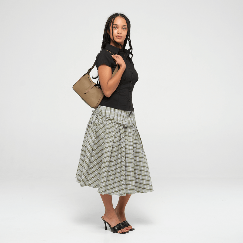 Woman holding an olive leather mini shoulder bag with a strap on a white background wearing a black top and plaid skirt on a white background
