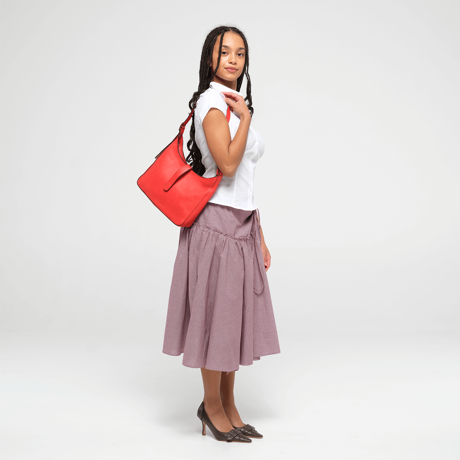 Woman holding a red leather shoulder bag  and wearing a white top and purple skirt on a white background