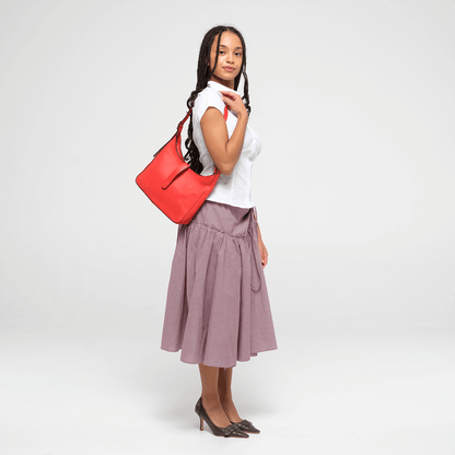 Woman holding a red leather shoulder bag  and wearing a white top and purple skirt on a white background